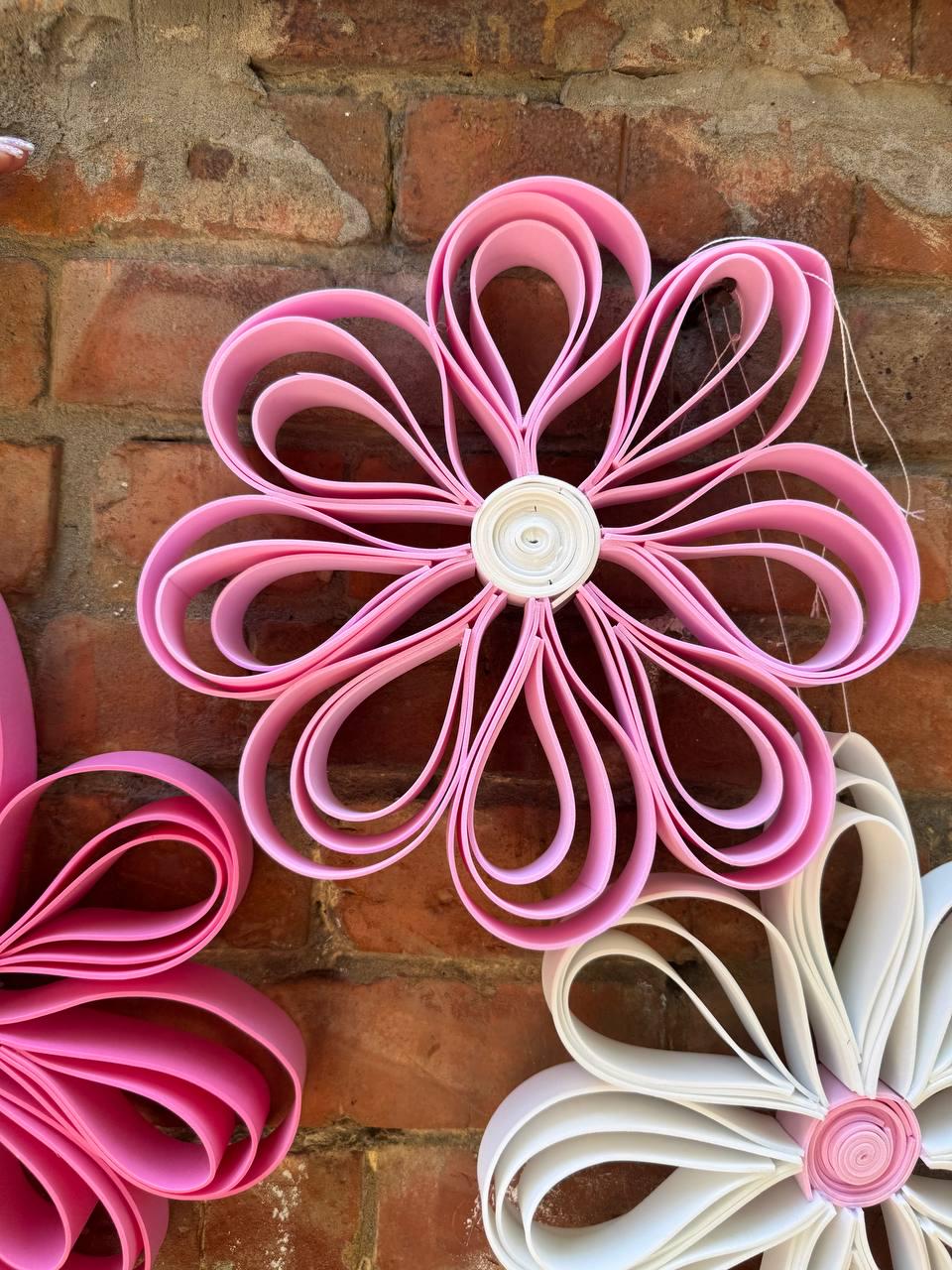 May include: A decorative wall hanging featuring a large flower made of folded pink paper strips arranged in a circular pattern with a white coiled paper center. The flower has multiple layered petals creating a three-dimensional effect.