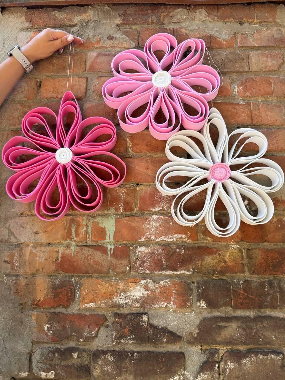 May include: Three hanging flower decorations made of looped strips. One flower is dark pink with a white center, one is light pink with a white center, and one is white with a light pink center. Each flower features eight petals formed by loops.