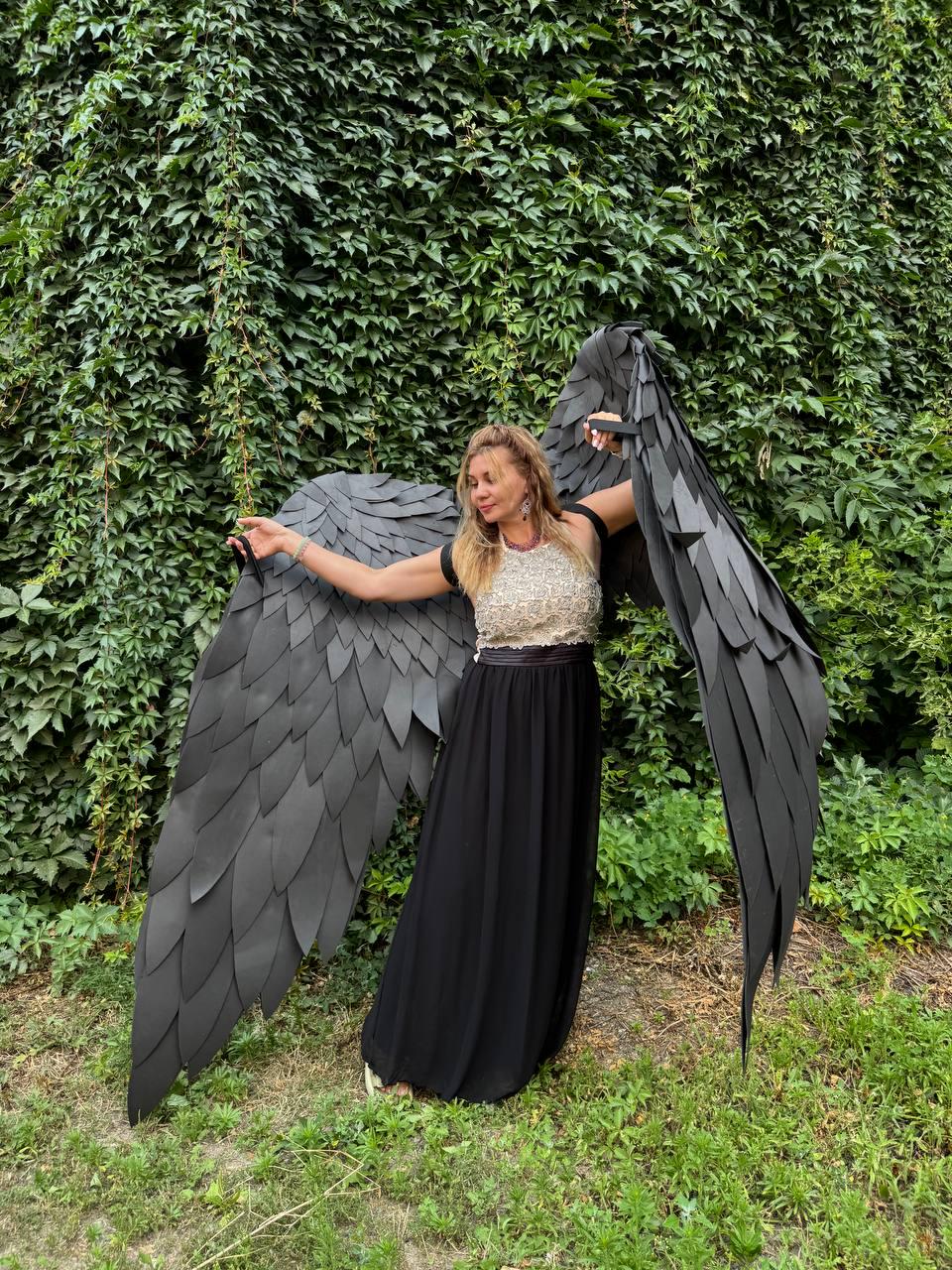May include: Large, articulated black wings constructed from layered, feather-shaped panels. The wings are designed to be worn on the back, featuring straps for attachment. The structure is lightweight and flexible, creating a realistic, expansive span.