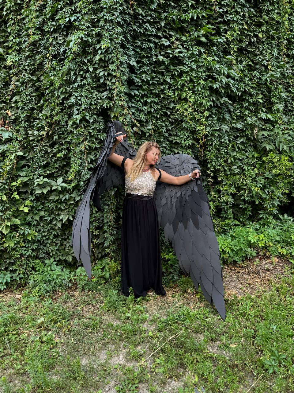 May include: A pair of large, black, layered feather-style wings attached to a garment featuring a sleeveless, textured, light-colored bodice and a long, flowing black skirt. The wings extend outward, showcasing a detailed, overlapping leaf-like pattern.