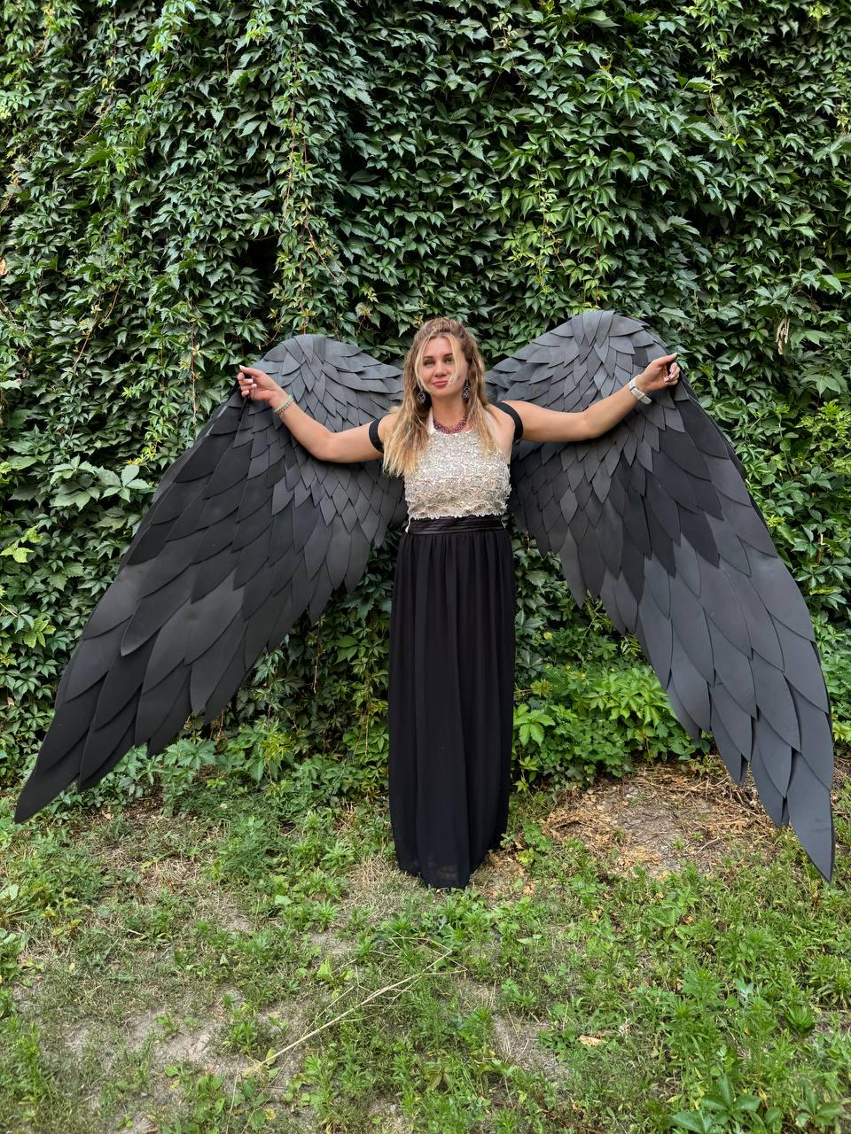 May include: A pair of large, black, articulated wings featuring layered, feather-like segments. The wings are designed to be worn on the back, extending outward with a wide span, showcasing a matte finish and a structured, pointed shape.
