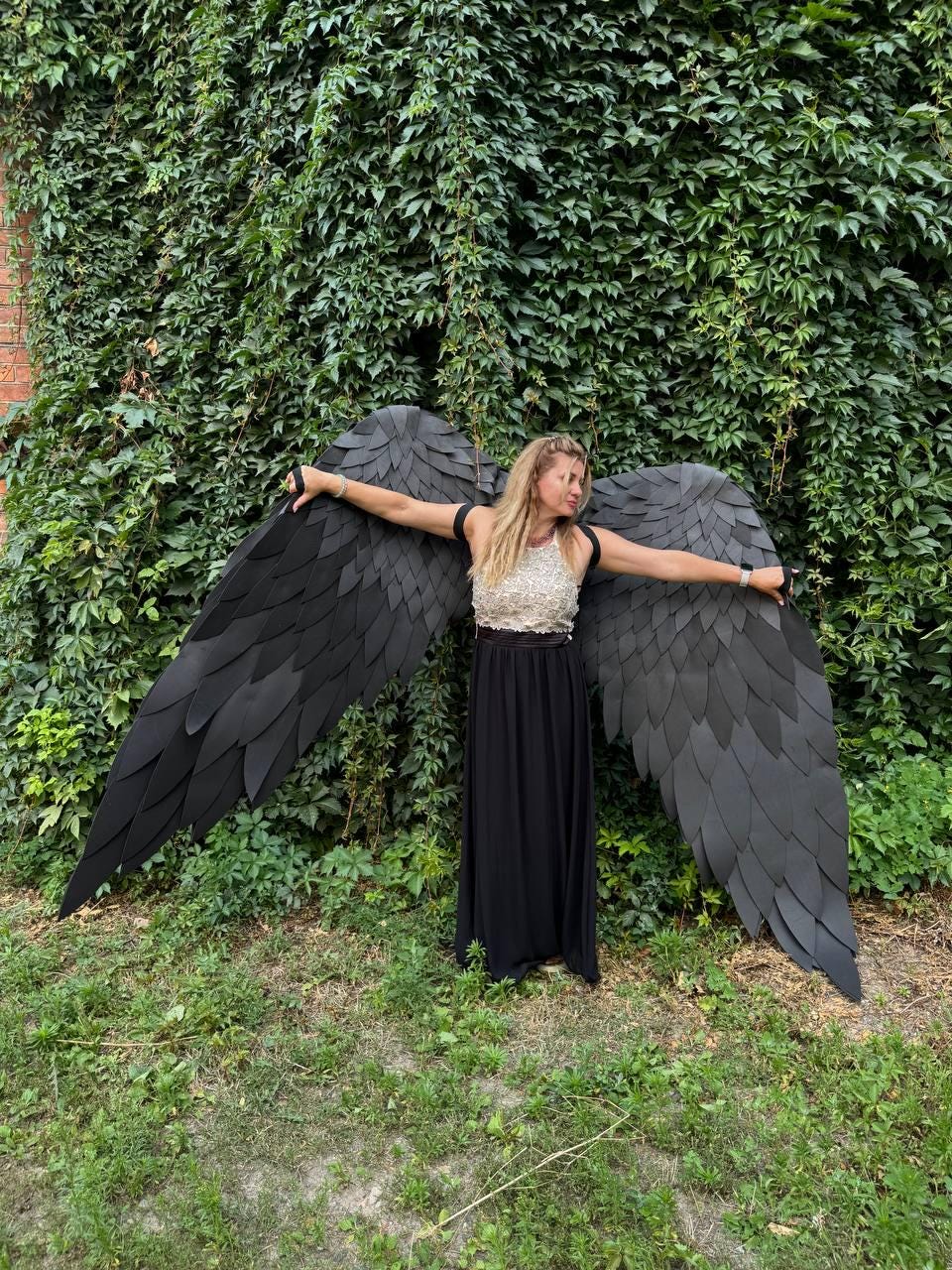 May include: Large black wings constructed from layered, feather-shaped panels. The wings are attached to a sleeveless top featuring a textured, light-colored bodice and a long, solid black skirt. The design creates a wide, expansive silhouette.