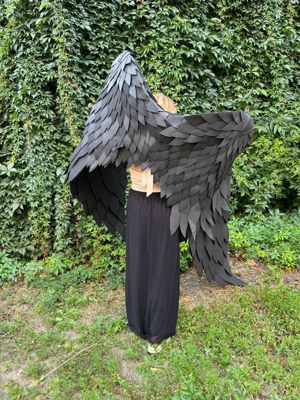 May include: Large black wings constructed from layered, feather-shaped pieces. The wings are worn over a beige top and a long black skirt. The design features a dense, overlapping pattern that mimics the appearance of bird plumage, extending outward from the center to create a wide, expansive shape that covers the upper back and shoulders.