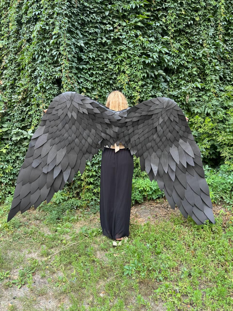 May include: A pair of large, black, wing-shaped accessories featuring layered, feather-like panels. The wings are wide and span horizontally, constructed from individual matte black segments that overlap to create a textured, realistic appearance.