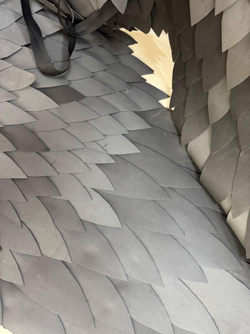 May include: A garment or accessory constructed from numerous overlapping, pointed, dark gray, leaf-shaped pieces arranged in a scale-like pattern. The structure includes a black strap visible at the top, creating a layered, textured appearance.