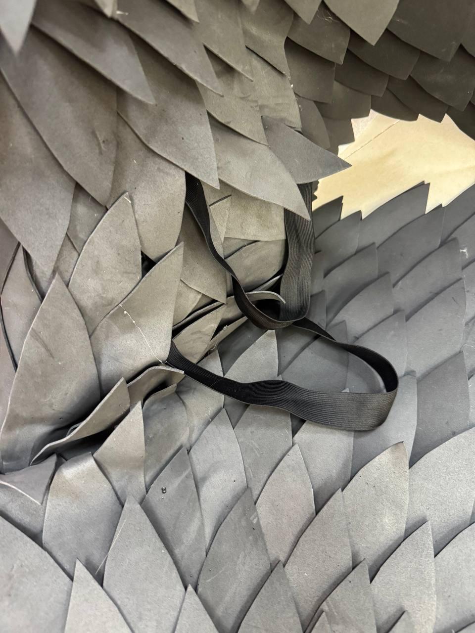 May include: A costume piece constructed from numerous overlapping grey foam scales arranged in a layered pattern. The structure features a flexible black elastic strap attached to the surface, allowing for adjustment and secure placement during use.