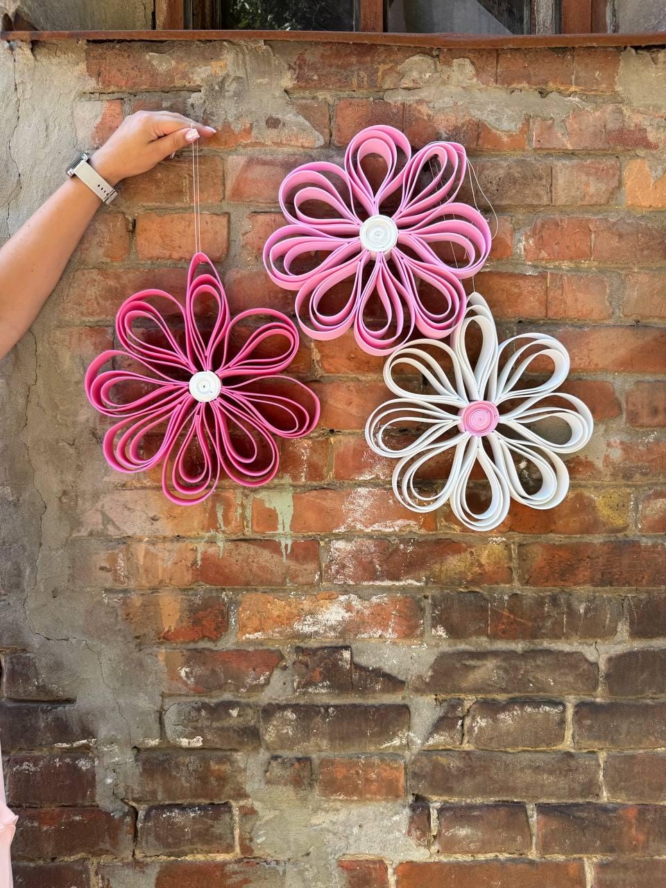May include: Three hanging flower ornaments constructed from looped strips of material. Two ornaments are vibrant pink, and one is white. Each flower features a central circular button-like accent. The petals are formed by folded loops arranged in a radial pattern.