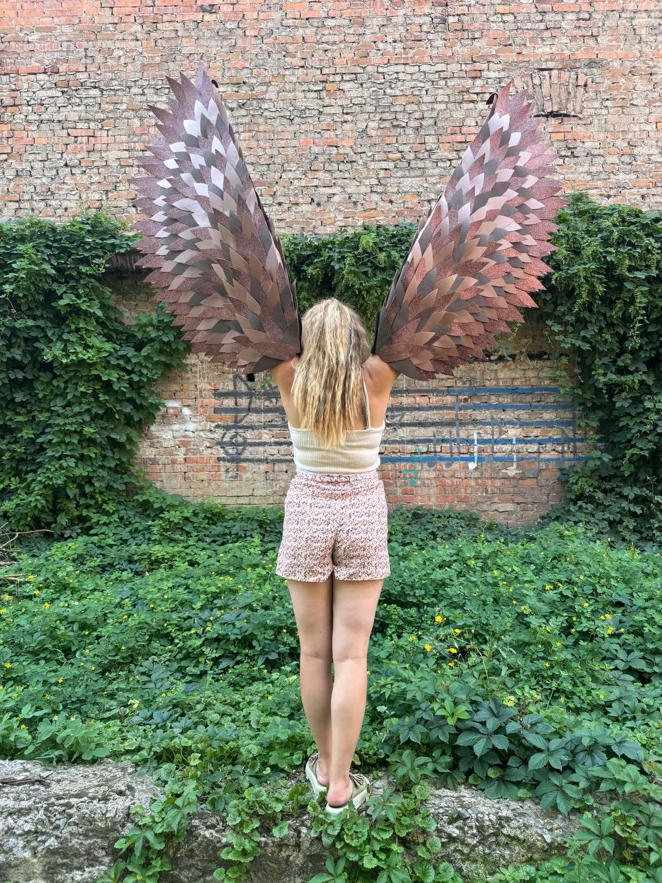May include: A pair of large, articulated wings featuring layered, pointed feathers in shades of brown and copper. The wings are constructed with a structured frame and display a gradient of dark to light tones, creating a realistic, expansive appearance.