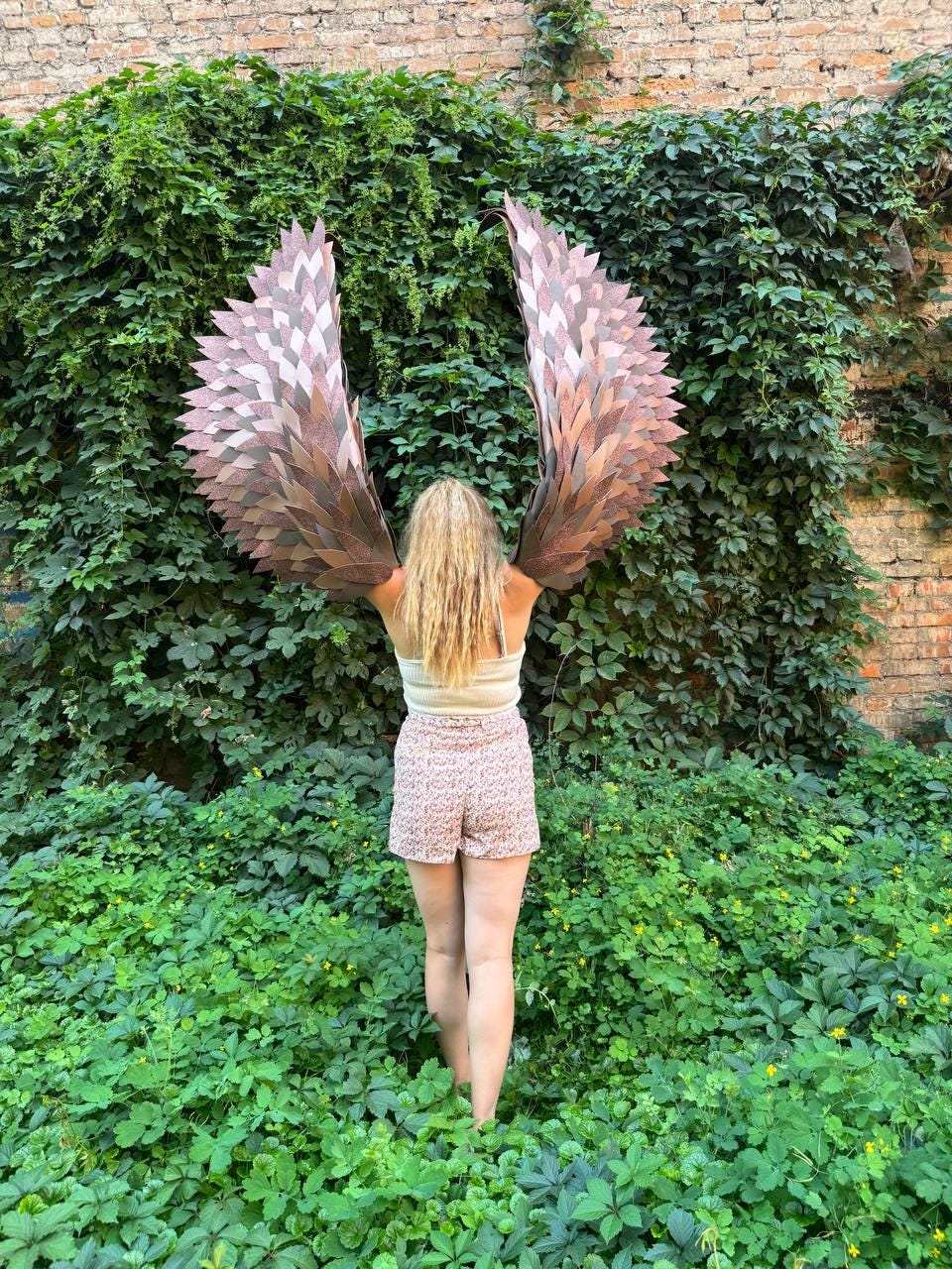 May include: A pair of large, decorative wings featuring layered, feather-like segments in shades of metallic copper and bronze. The wings are constructed with a pointed, angular design, extending upward and outward with a textured, overlapping finish.