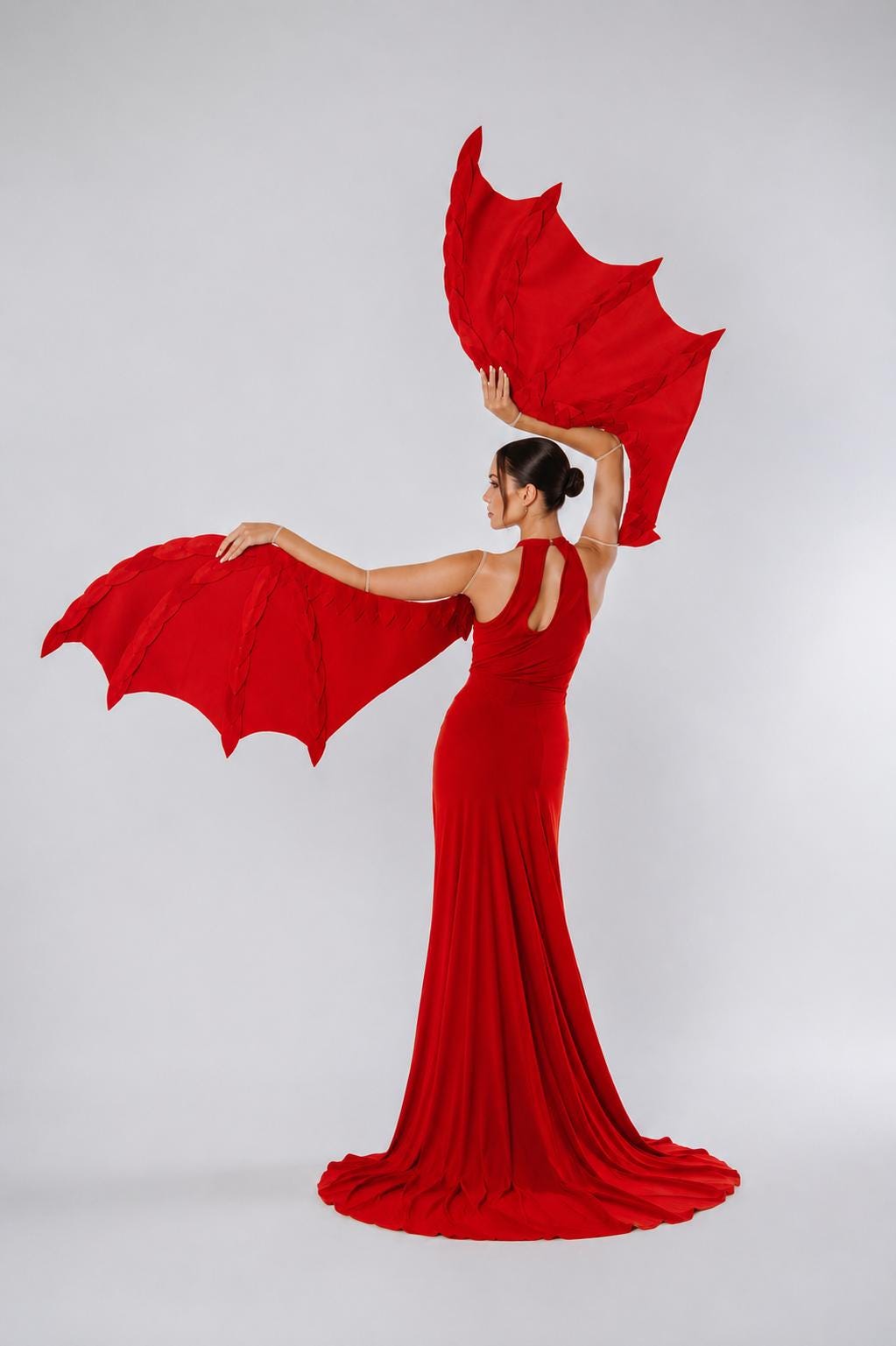May include: A floor-length red dress with a high neckline and a keyhole cutout on the back, paired with two large, articulated red wing-shaped accessories held by the arms, featuring a structured, webbed design with pointed edges.