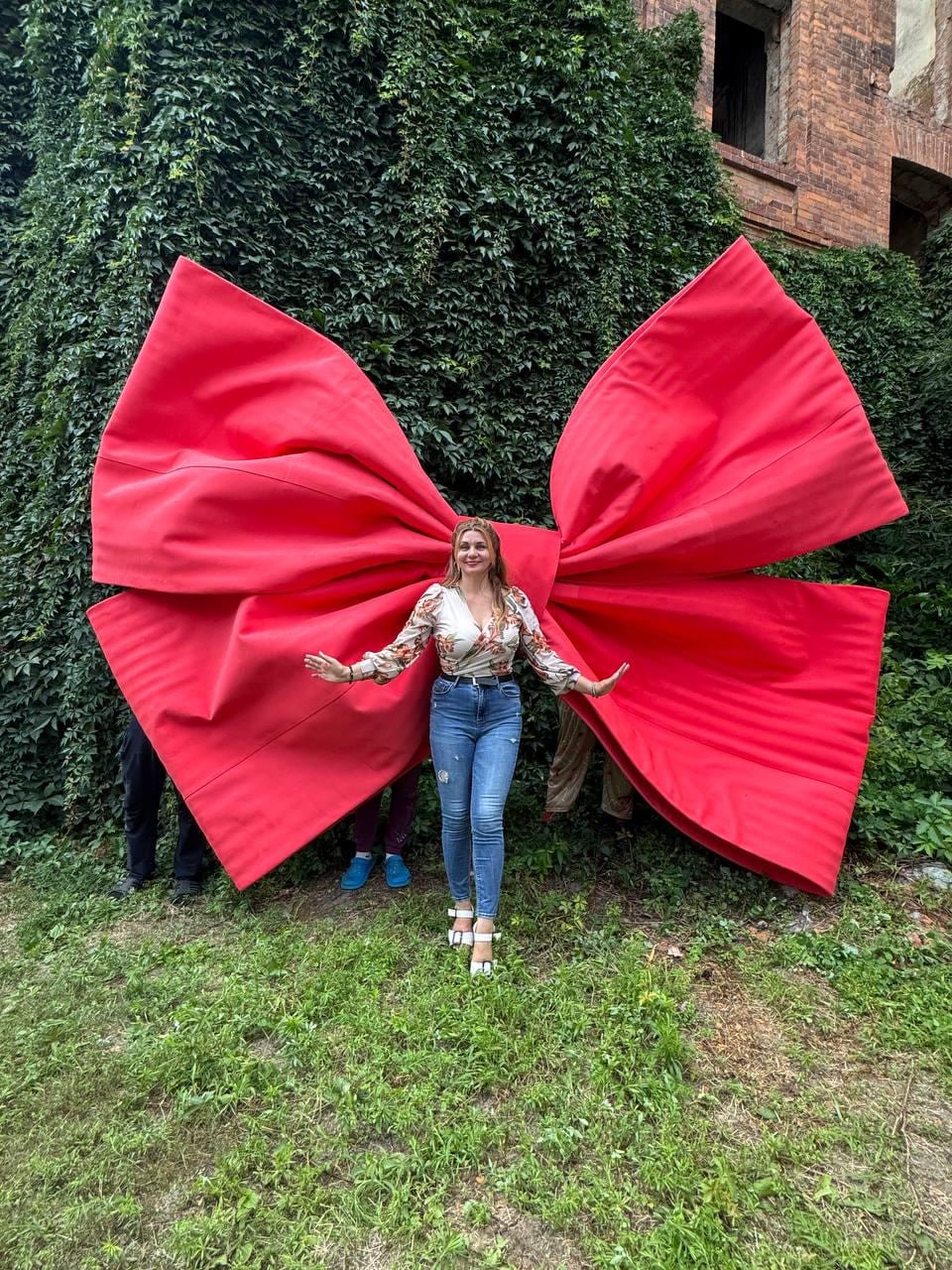 May include: A large, bright red decorative bow made of fabric, positioned behind a person wearing a long-sleeved floral print top, blue denim jeans, and white sandals. The bow features prominent folds and a central knot, standing against a green wall.