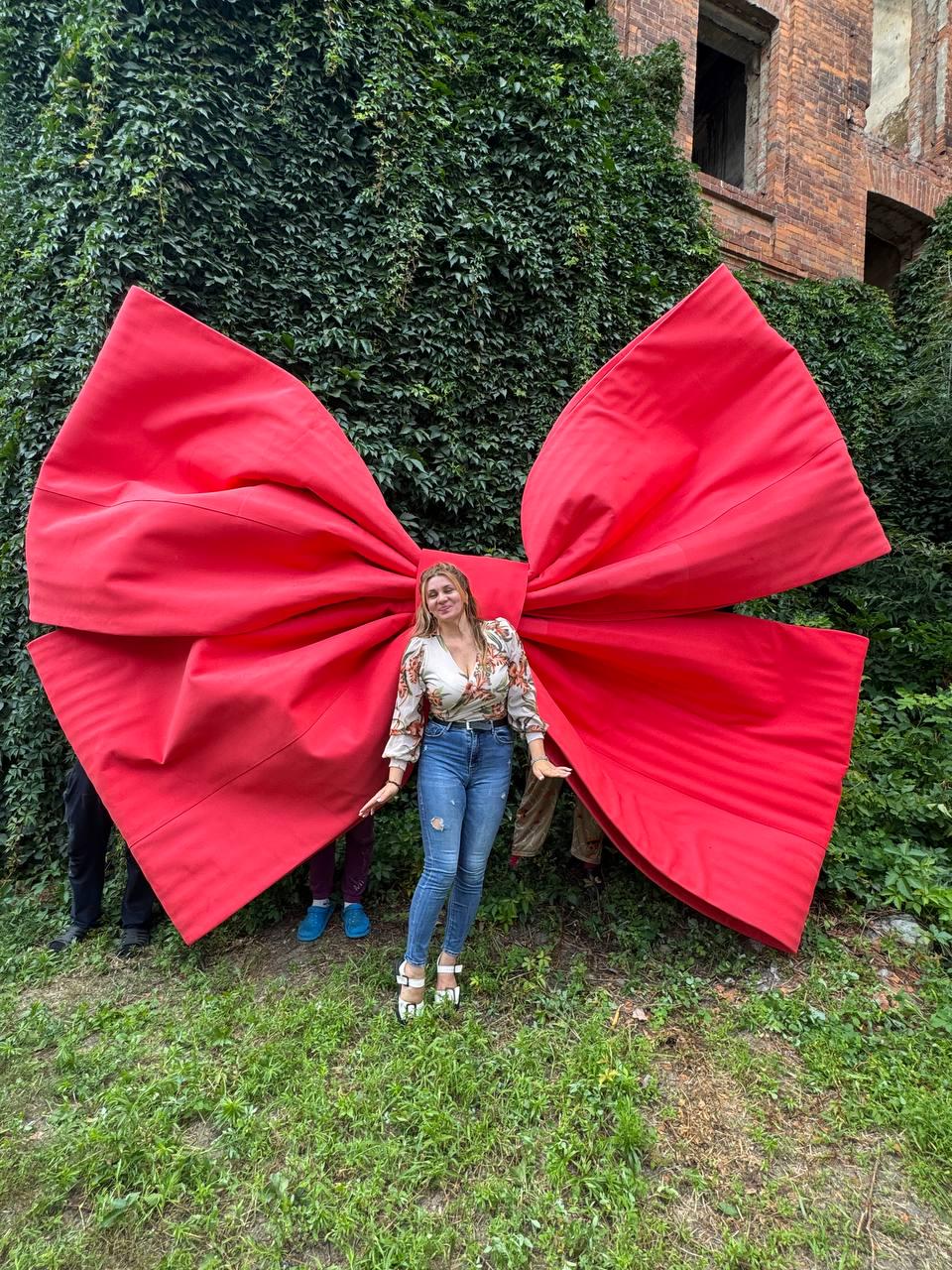 May include: A large, oversized red fabric bow with wide, structured loops and long, flowing tails. The bow is constructed from a solid, matte red material and features prominent, deep folds throughout its surface, creating a voluminous appearance.