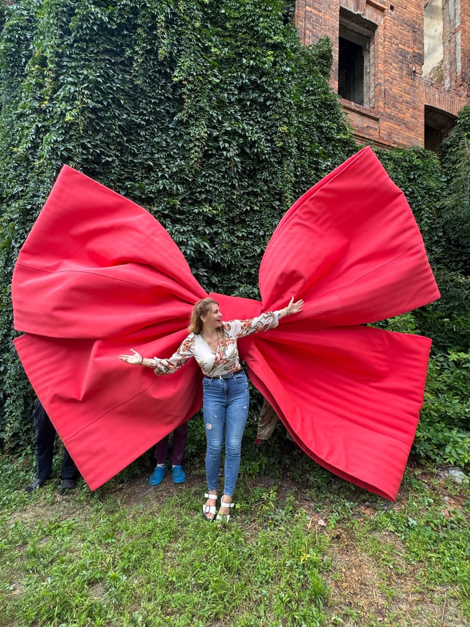 May include: A giant, oversized red fabric bow with wide, structured loops and long, flowing tails. The bow is constructed from a smooth, solid red material and features prominent, straight stitched seams running horizontally across the fabric surface.