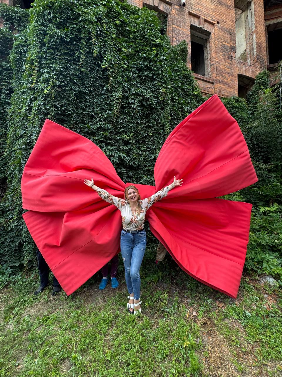 May include: A large, oversized red fabric bow with wide, structured loops. The bow is positioned behind a person wearing a long-sleeved floral print top, blue denim jeans, and white sandals, creating the appearance of wings or a decorative backdrop.