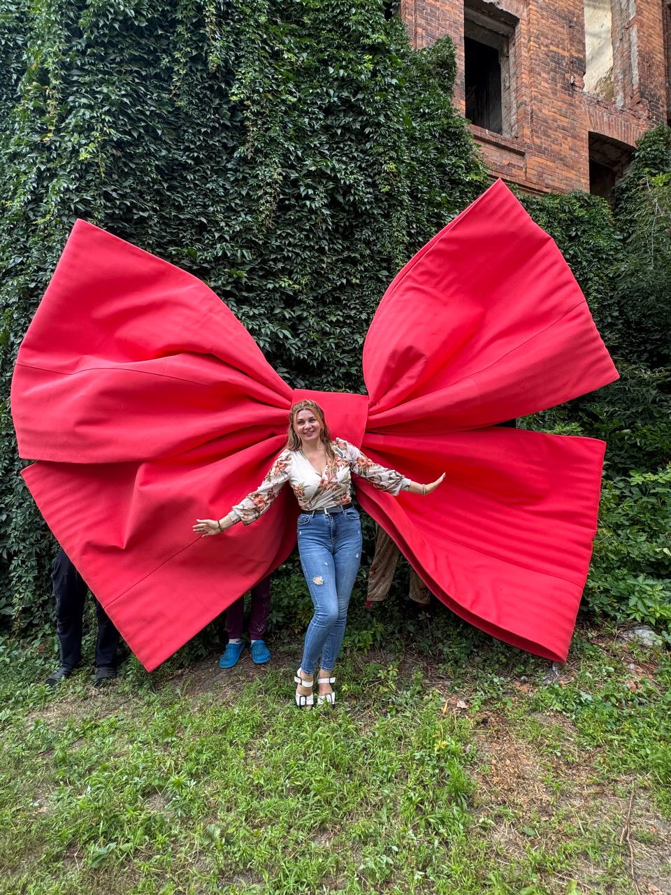 May include: A massive, oversized bright red fabric bow with structured, pleated wings. The bow is positioned behind a person wearing a white floral print long-sleeve wrap top, blue denim jeans, and white platform sandals, standing in front of greenery.