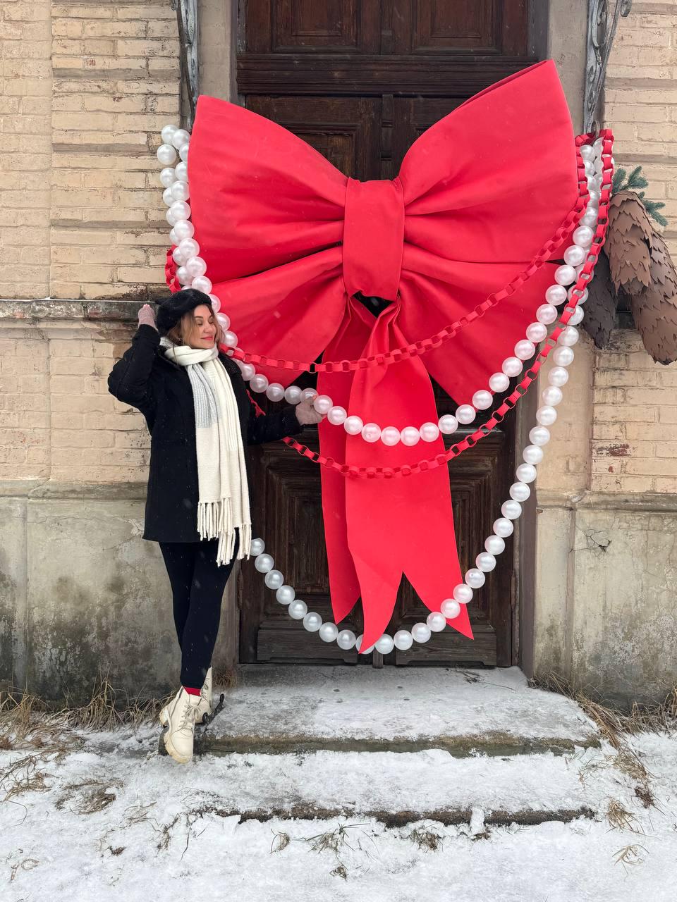 May include: A large red fabric bow decoration mounted on a dark wooden door, adorned with long strands of white spherical beads and red chain links. The bow features two wide loops and two long, pointed tails hanging down the center of the door.