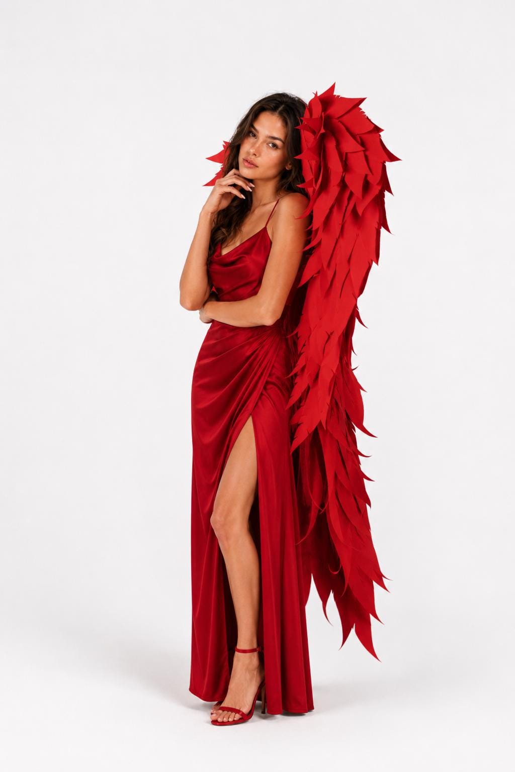 May include: A floor-length red satin dress with a cowl neckline, thin spaghetti straps, and a high side slit, paired with large, structured red wings featuring layered, pointed feather-like segments and matching red high-heeled sandals.