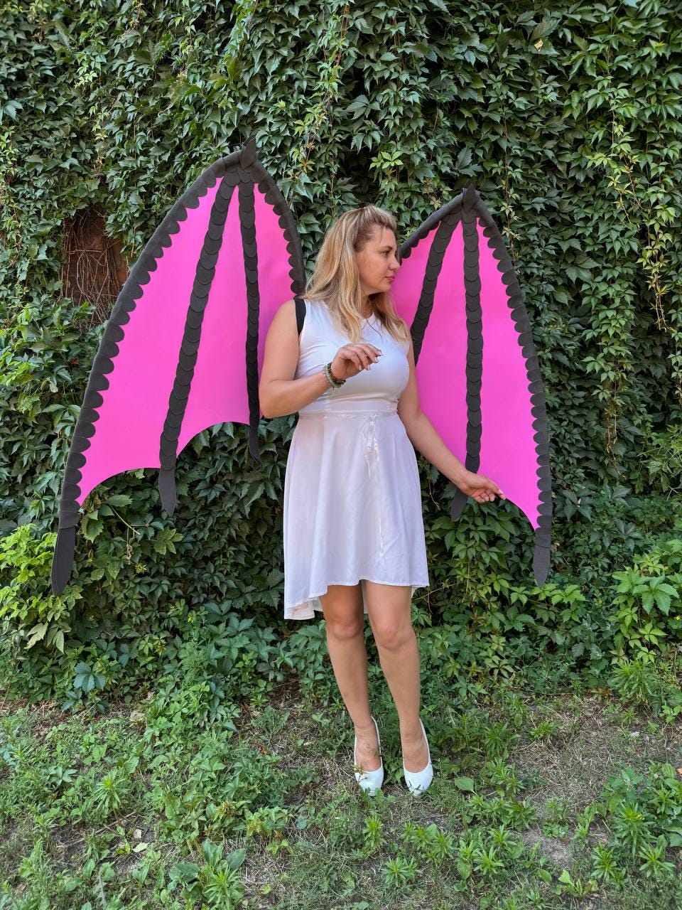 May include: A white sleeveless dress paired with large, detachable dragon wings. The wings feature bright pink panels with black scalloped edges and structural ribs, worn over the shoulders with black straps against a green leafy background.