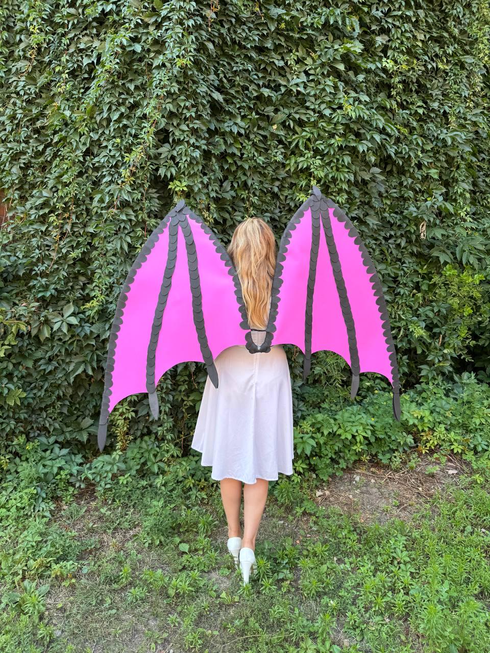 May include: A pair of large, bright pink dragon wings with black scalloped edges and structural ribs, worn over a simple white dress. The wings feature a pointed, bat-like shape with distinct segments and a central harness attachment point.