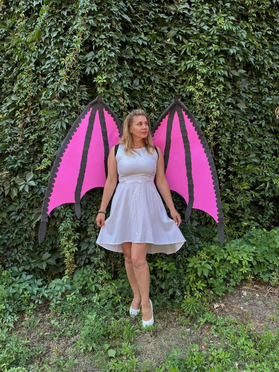 May include: A sleeveless white knee-length dress paired with large, detachable dragon-style wings. The wings feature vibrant pink panels framed by black scalloped edges and structural ribs, worn like a backpack over the shoulders of the dress.