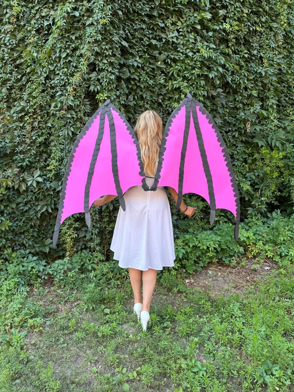May include: Large dragon-style wings featuring vibrant pink panels with black structural ribs and scalloped edges. The wings are attached to a central harness worn on the back, extending outward with a pointed, angular shape and a distinct bat-like design.