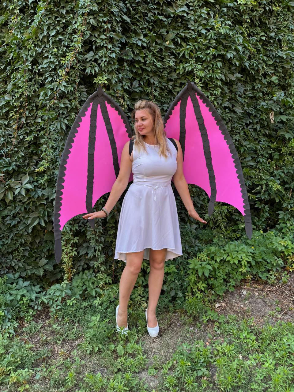 May include: A white sleeveless dress with a flared skirt paired with large, detachable bat-style wings. The wings feature bright pink panels framed by thick black borders with scalloped edges, secured by black straps worn over the shoulders.