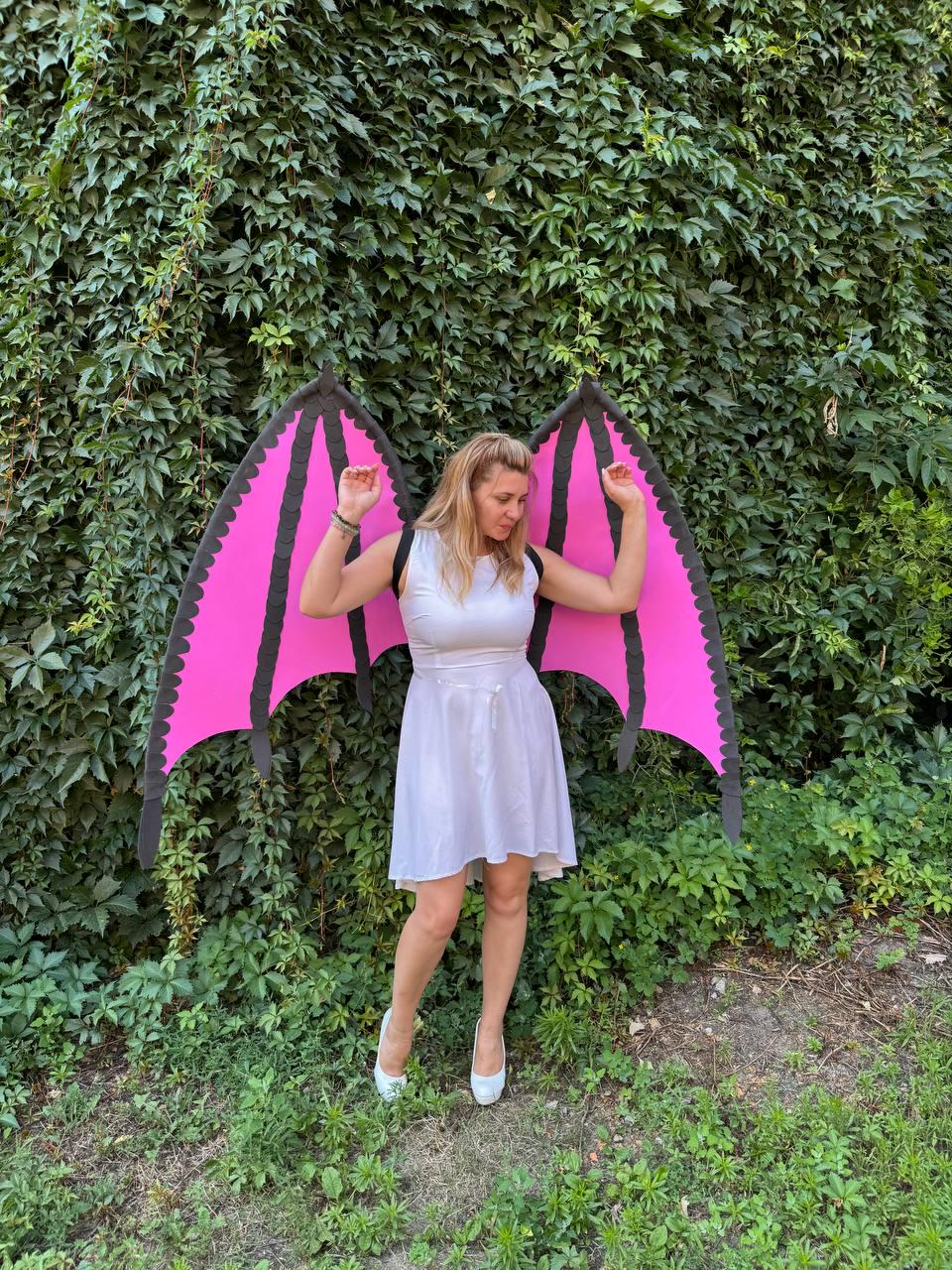 May include: A white sleeveless dress paired with large, detachable bat-style wings. The wings feature a vibrant pink interior with black structural framing and scalloped edges. The dress has a fitted bodice and a flared skirt that reaches above the knee.