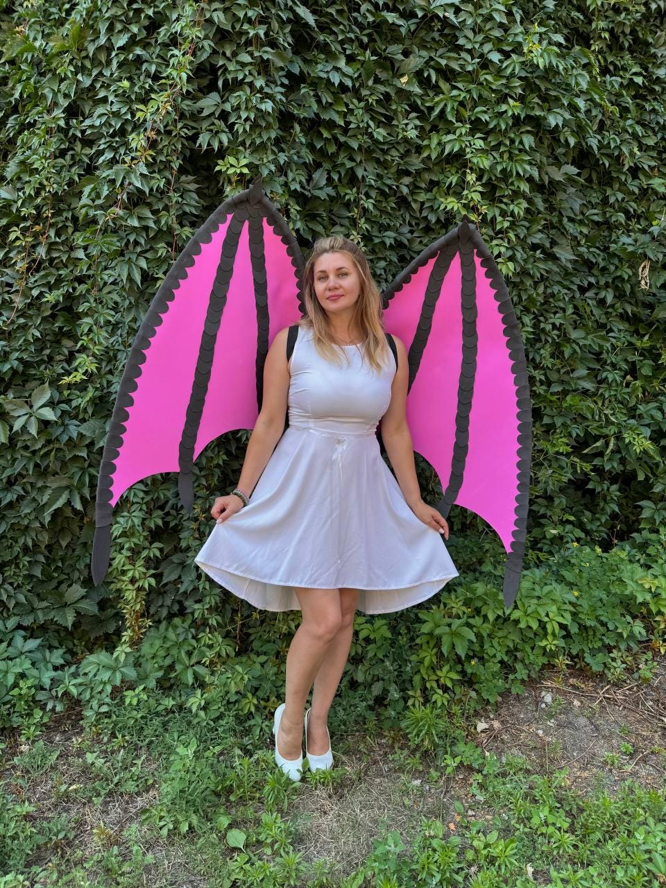 May include: A white sleeveless dress with a flared skirt paired with large, detachable bat-like wings. The wings feature bright pink panels framed by thick black borders with scalloped edges, secured by black straps worn over the shoulders.