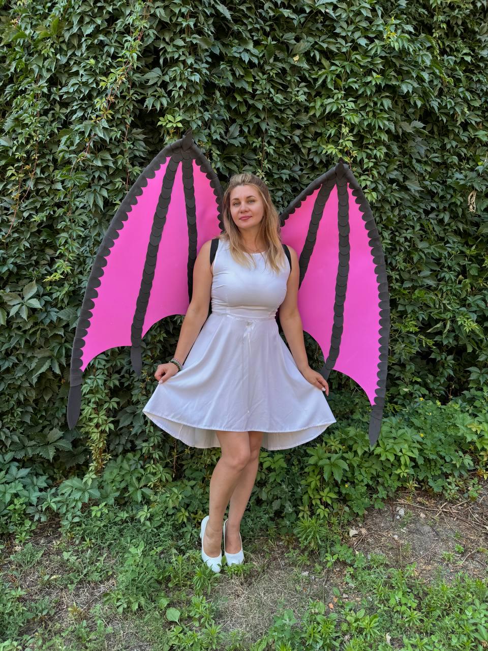 May include: A white sleeveless dress with a flared skirt paired with large, detachable bat-like wings. The wings feature bright pink panels framed by thick black borders with scalloped edges, secured by black straps worn over the shoulders.