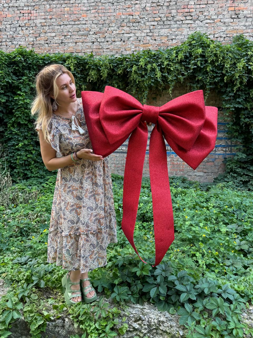 May include: A large, oversized decorative bow in a vibrant, glittery red finish. The bow features two wide, long ribbon tails hanging downward from the center knot, with two expansive, structured loops on each side, creating a bold, festive appearance.