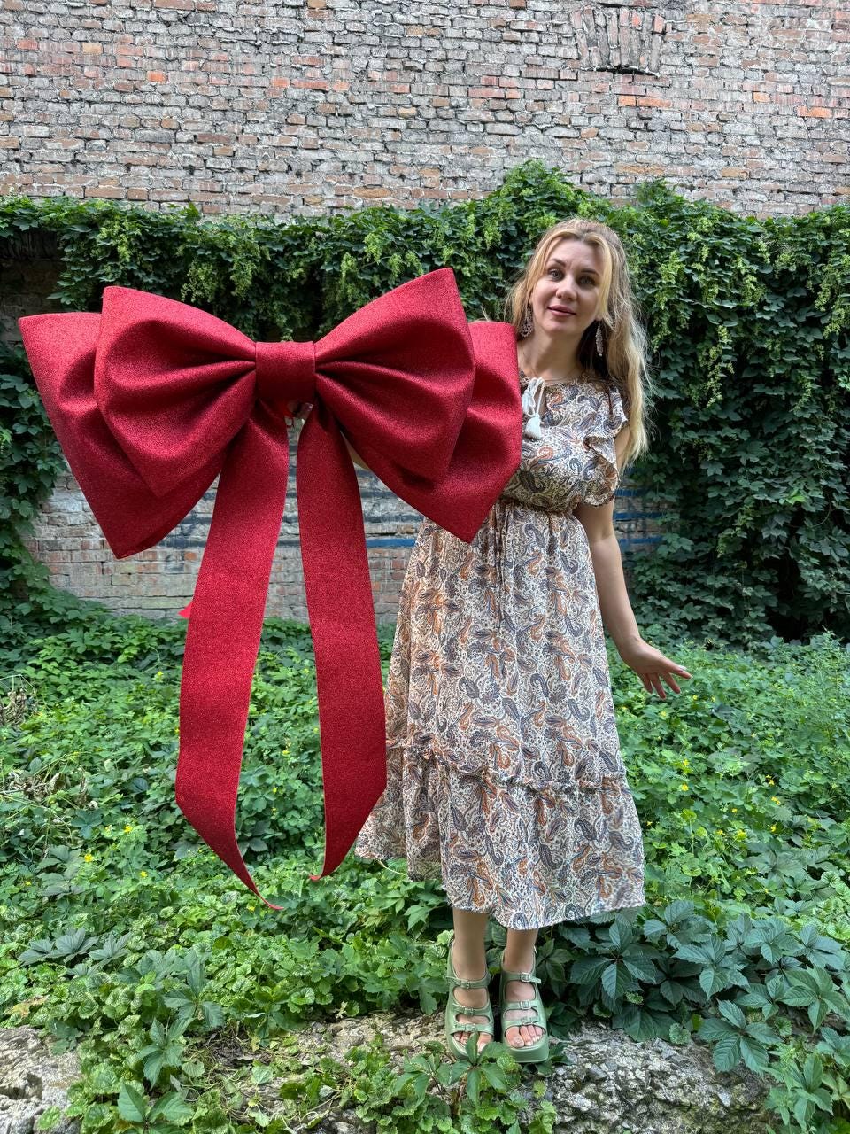 May include: A large, glittery red decorative bow featuring two wide, long hanging tails. The bow has a textured, sparkling surface and is constructed with multiple loops to create a voluminous, three-dimensional shape for display purposes.