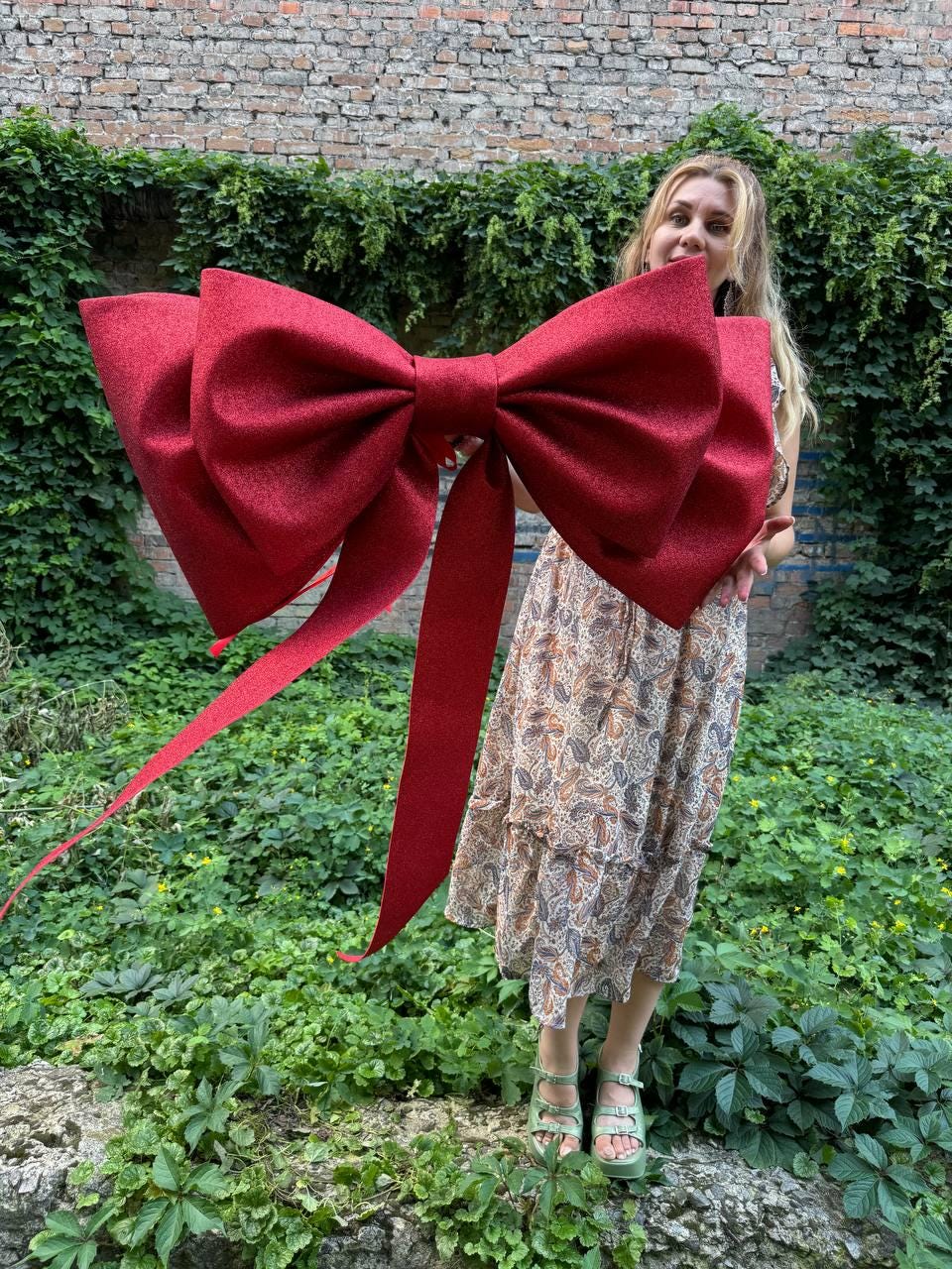 May include: A large, oversized decorative bow in a deep, shimmering red color. The bow features a textured, glittery finish with two prominent loops and two long, trailing ribbon tails hanging downward from the center knot of the bow structure.