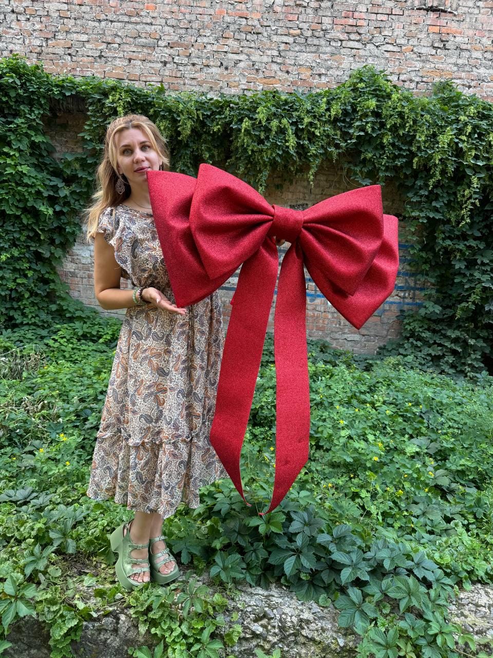 May include: A large, oversized decorative bow in a deep red color with a prominent glittery texture. The bow features two wide, structured loops and two long, trailing ribbon tails that hang vertically, creating a bold and festive appearance.