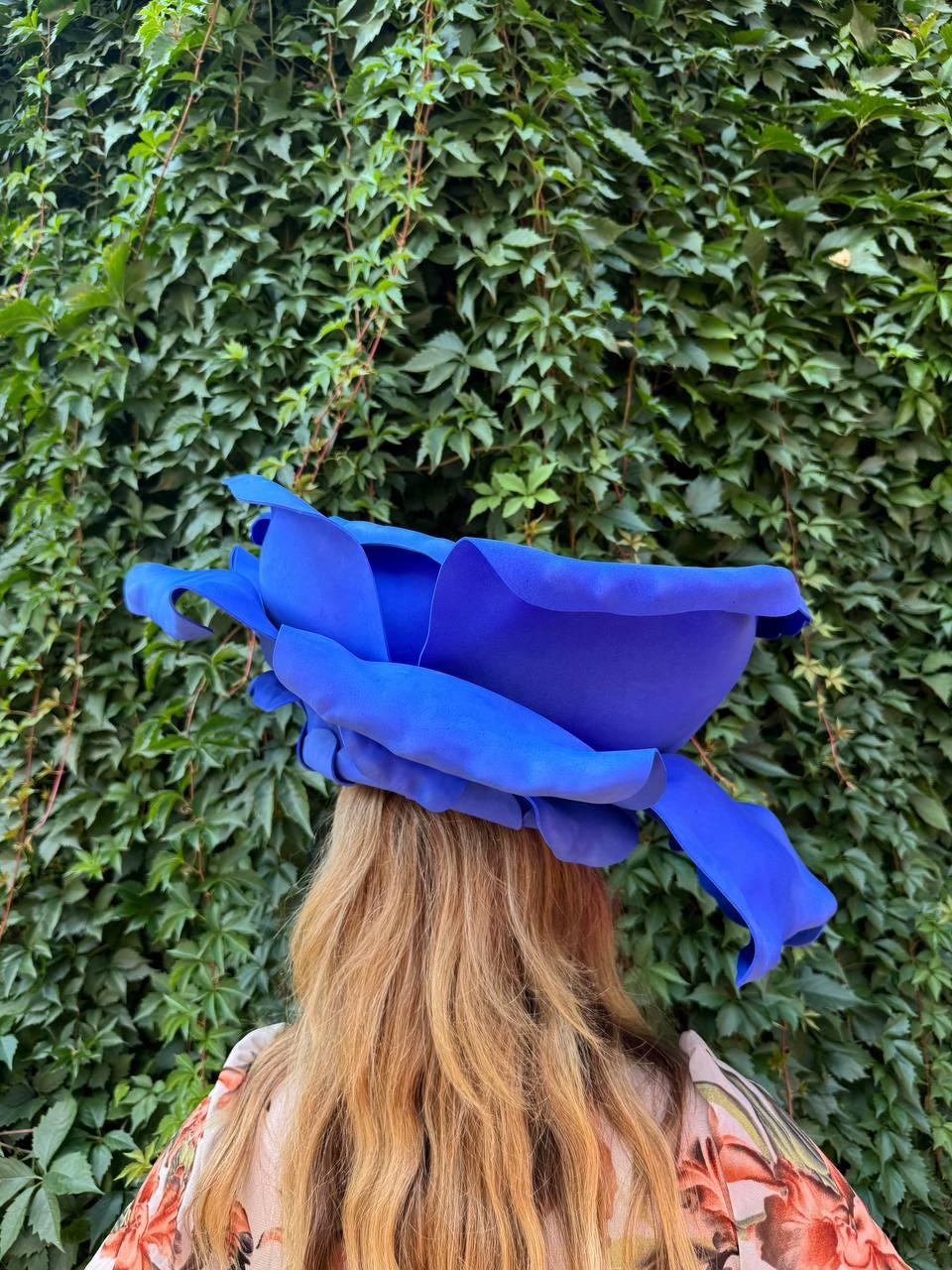 May include: A large, vibrant blue hat shaped like an oversized flower with layered, curved petals. The hat features a deep, saturated blue color and a sculptural design that mimics the appearance of a blooming blossom, resting atop long hair.