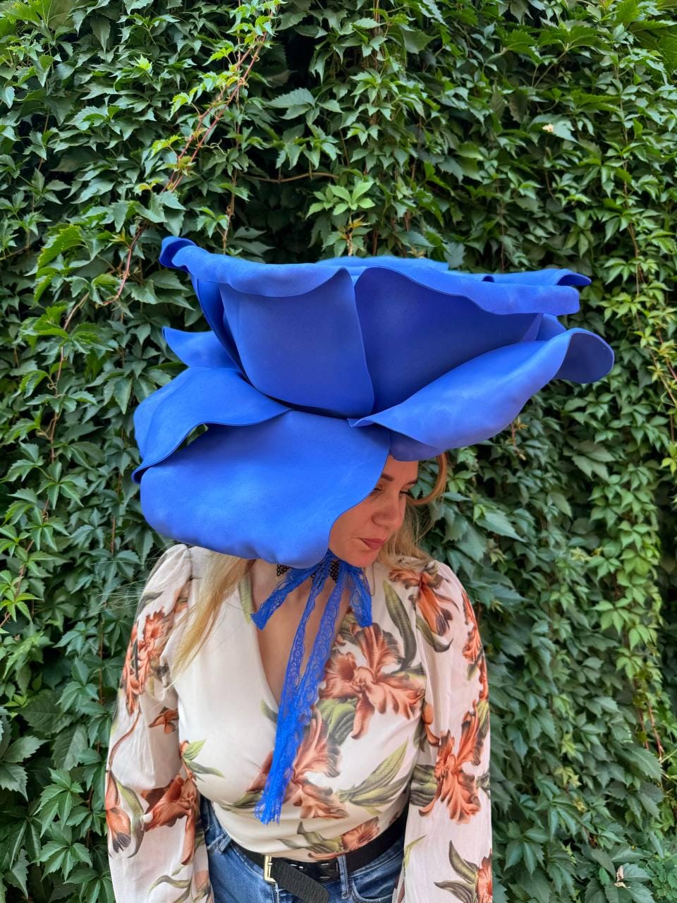 May include: A large, oversized blue rose headpiece featuring wide, sculpted petals. The accessory is secured with a thin, blue lace ribbon tied in a bow under the chin, creating a bold, floral-inspired statement piece for the head.
