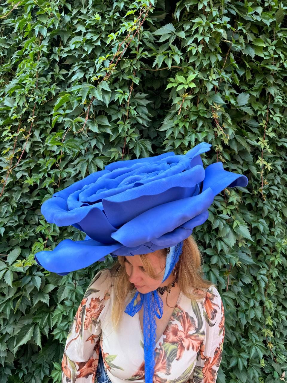 May include: A large, oversized blue rose headpiece featuring layered, sculpted petals. The accessory is secured with a blue lace ribbon that ties under the chin, complementing the floral design with its vibrant color and intricate, structured form.