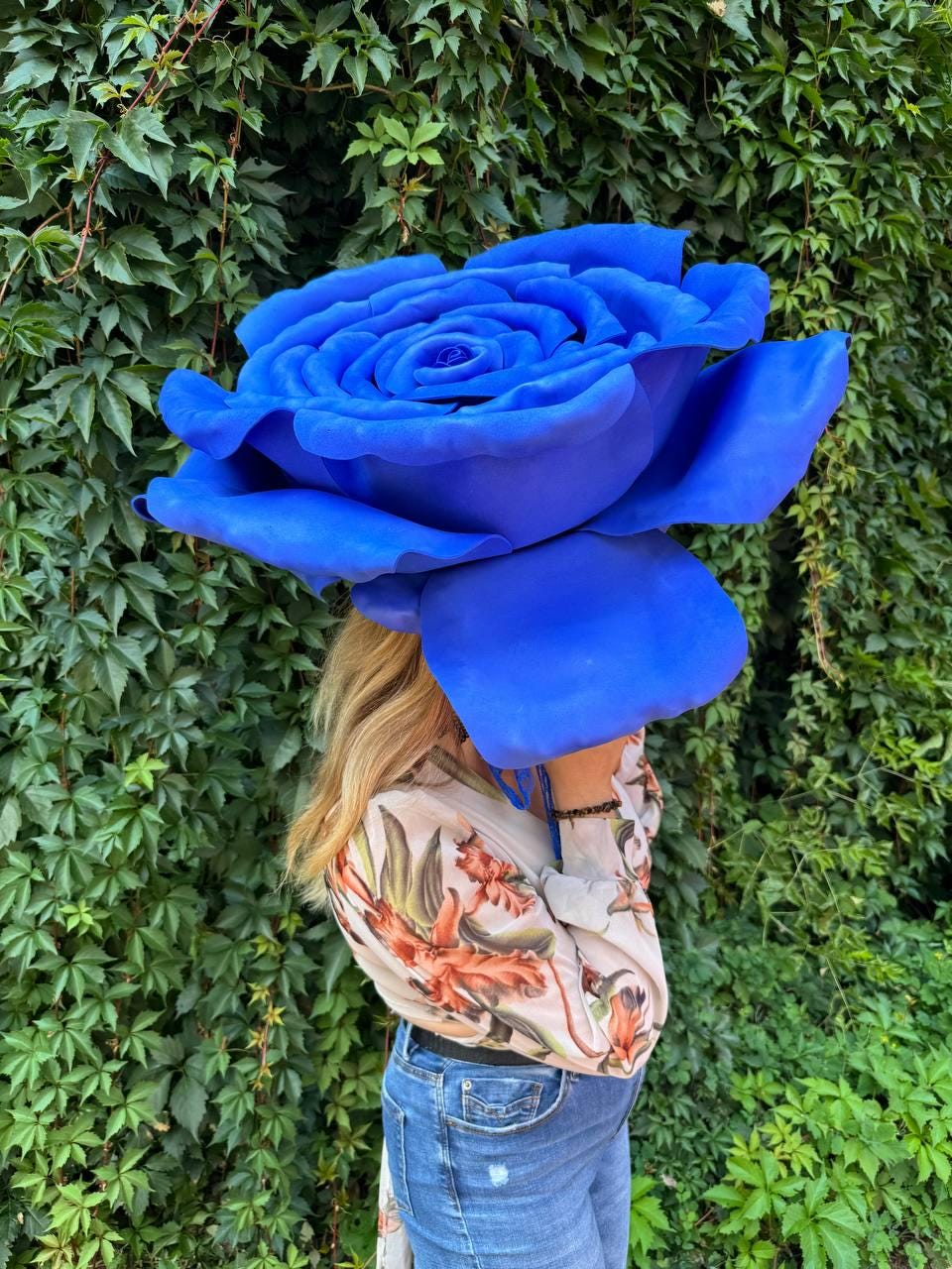 May include: A large, oversized artificial blue rose with layered, rounded petals. The flower is held in front of the face, obscuring it. The rose features a vibrant, solid blue color throughout its structure, with a dense, multi-layered petal arrangement.