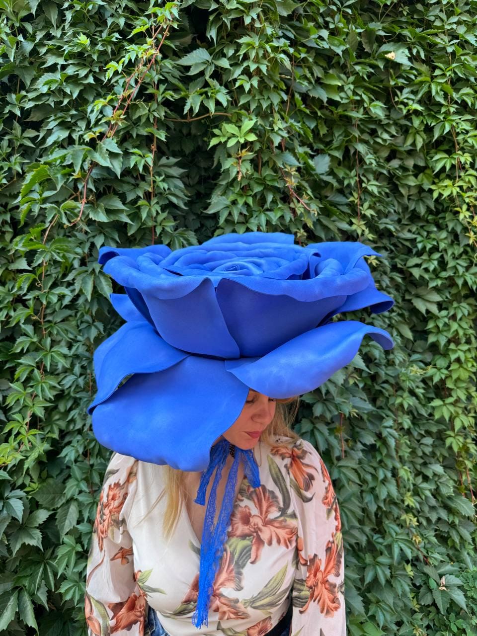May include: A large, oversized blue rose headpiece featuring layered, sculpted petals. The accessory includes a thin, blue lace ribbon tie that hangs down from the center. It is worn with a long-sleeved top featuring a cream base and orange floral print.