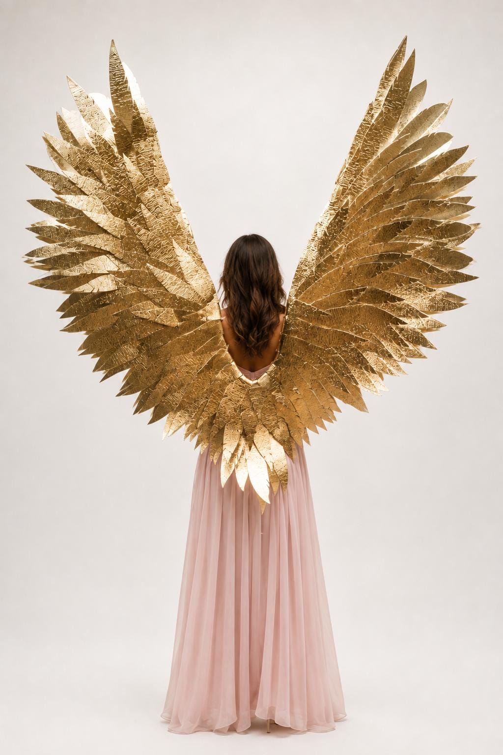 May include: Large, metallic gold wings featuring a layered, textured feather design. The wings are attached to a long, floor-length, pale pink dress with a flowing skirt. The wings extend upward and outward, showcasing a bright, reflective finish.