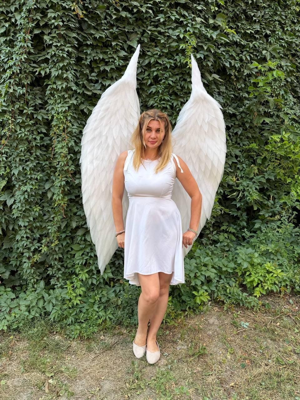 May include: A white sleeveless dress with a flared skirt paired with large, feathered white wings. The wings are tall and pointed, extending upwards behind the shoulders, featuring layered white plumage that creates a textured, expansive appearance.