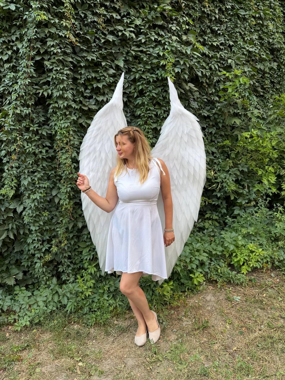 May include: A pair of large, white, feathered angel wings attached to a white sleeveless dress. The wings feature layered, pointed feathers that extend upward and outward, creating a dramatic silhouette behind the wearer of the white dress.