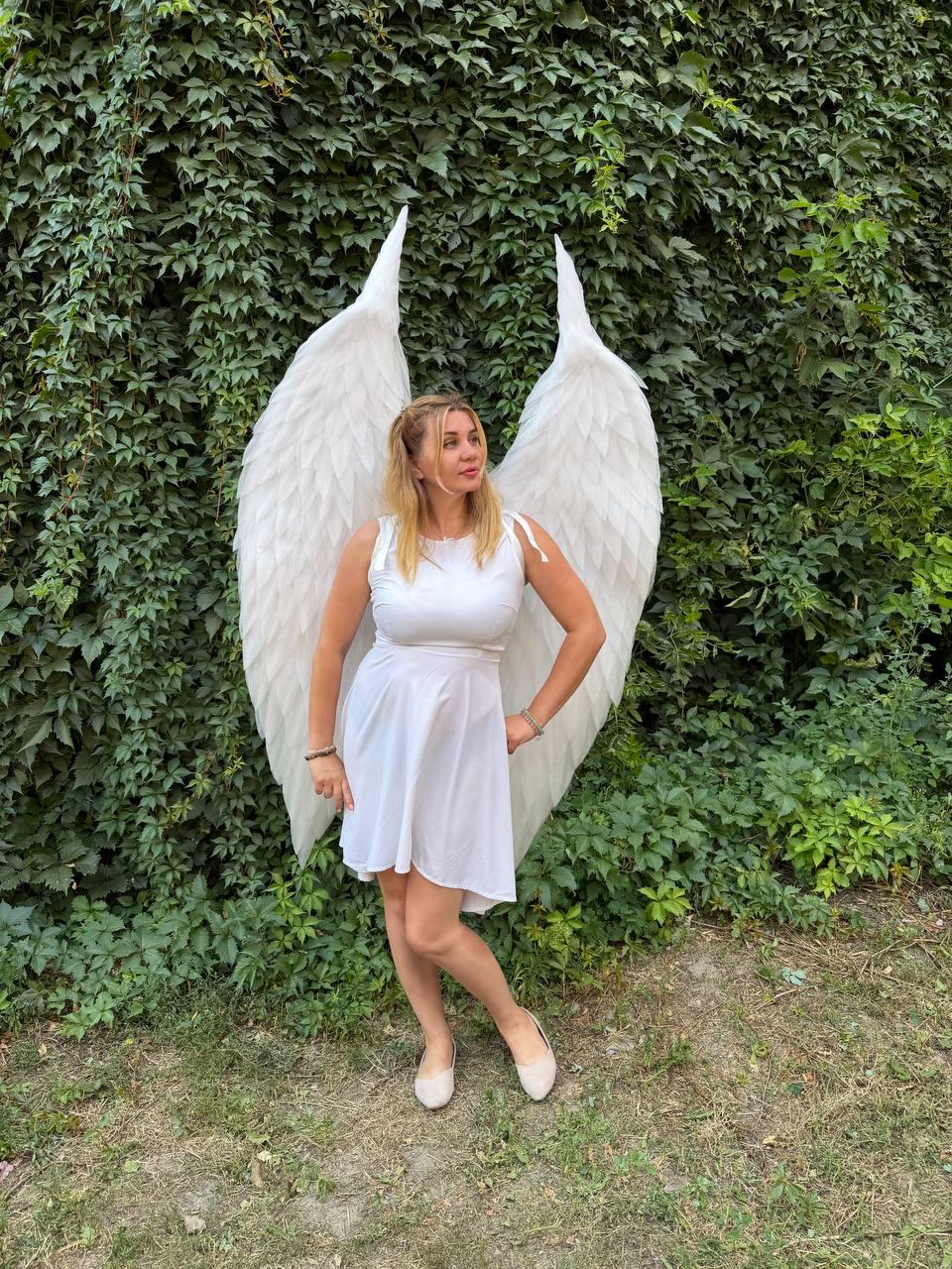 May include: A white sleeveless dress paired with large, feathered white angel wings. The dress features a fitted bodice and a flared skirt that reaches above the knees. The wings are tall, pointed, and extend upwards, creating a dramatic silhouette.