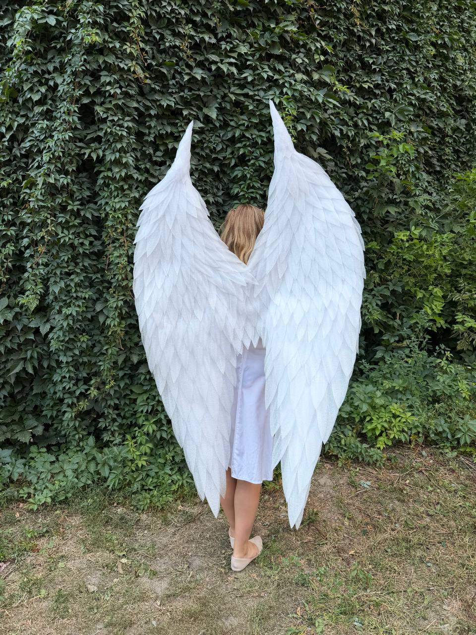 May include: Large, white, feathered angel wings designed to be worn on the back. The wings feature layered, pointed white feathers that extend upward and outward, creating a full, expansive shape that mimics the appearance of bird wings.