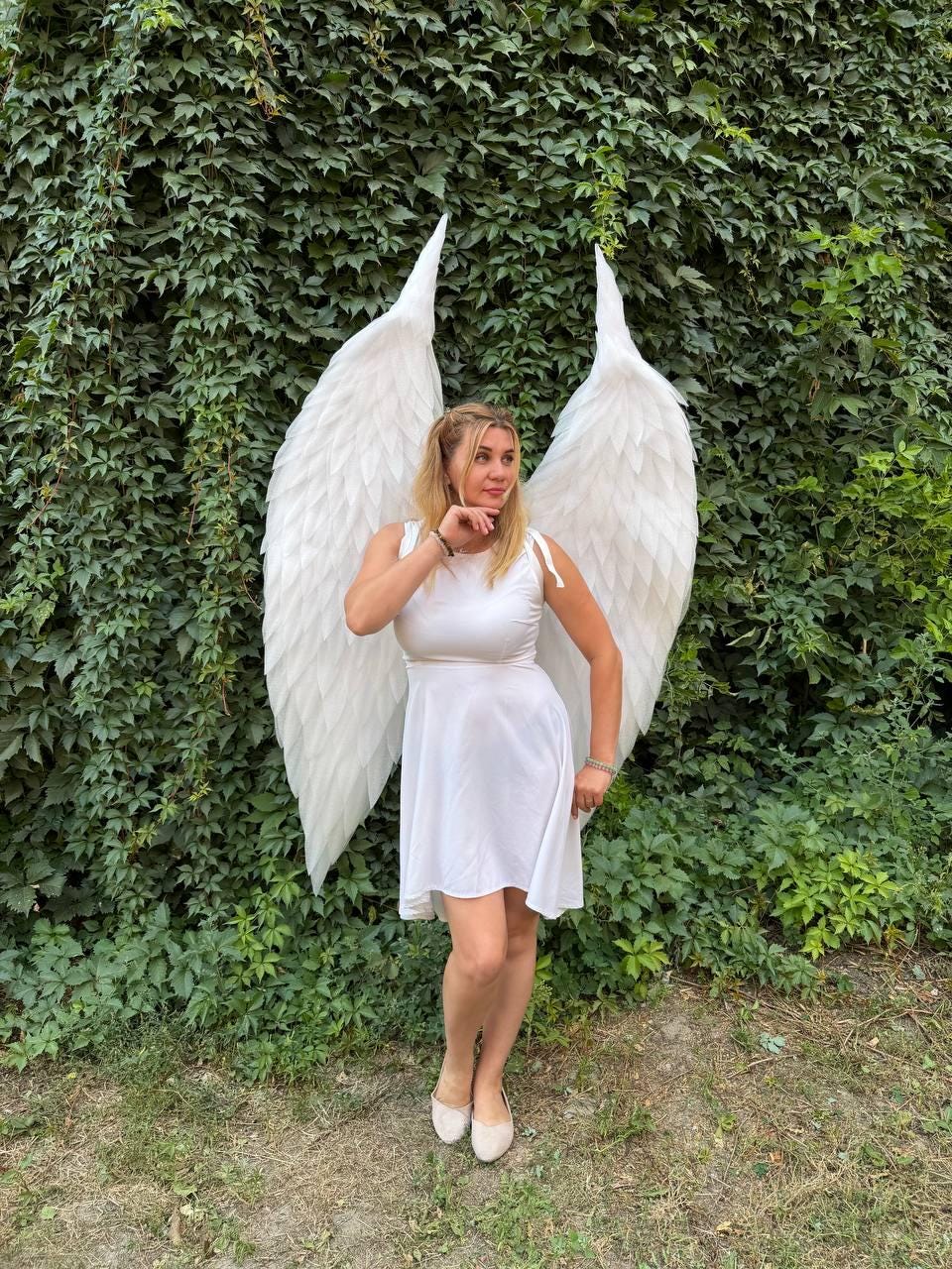 May include: A pair of large, white, feathered angel wings attached to a white sleeveless dress. The wings feature layered, pointed feathers that extend upward and outward, creating a dramatic span behind the white dress with a scoop neckline.