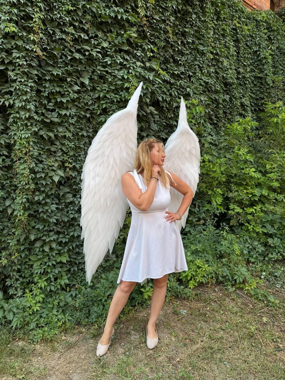 May include: A pair of large, white, feathered angel wings attached to a white sleeveless dress. The wings feature layered, pointed white feathers that extend upward and outward, creating a dramatic silhouette behind the white dress.