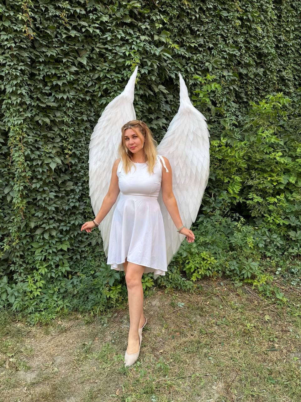 May include: A white sleeveless dress with a flared skirt and a pair of large, white, feathered-style wings attached to the back. The wings feature layered, pointed segments that extend upward and outward, creating a wide, expansive silhouette.