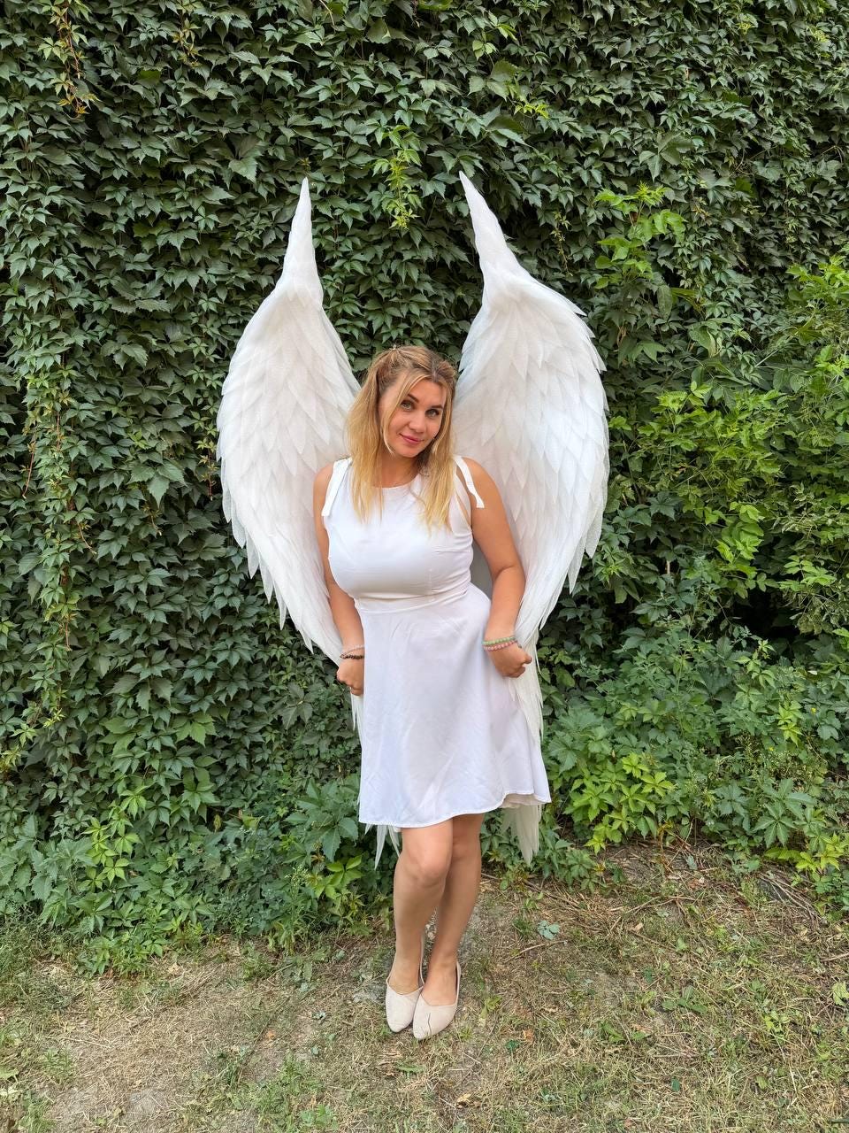 May include: A white sleeveless dress with a round neckline and a pair of large, tall, white feathered wings attached to the back. The wings feature layered, pointed feathers that extend upward and outward, creating a dramatic, expansive silhouette.