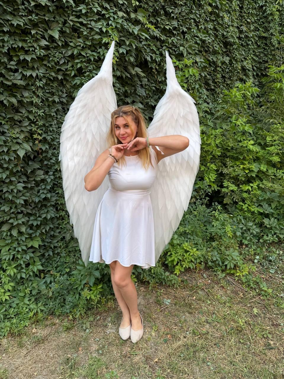 May include: A white sleeveless dress paired with large, feathered white angel wings. The dress features a fitted bodice and a flared skirt that reaches above the knees. The wings are tall, pointed, and constructed with layered white feathers.