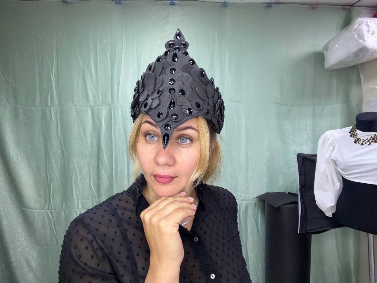 May include: A black headpiece featuring a pointed crown structure composed of layered, petal-like shapes. The surface is adorned with numerous glossy, black teardrop-shaped crystals arranged in vertical patterns along the center and sides.