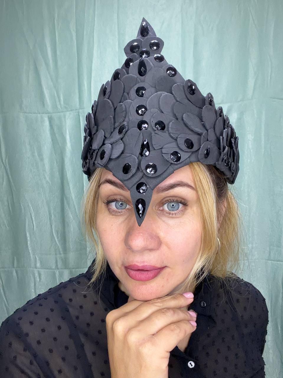 May include: A pointed headpiece constructed from layered matte black petal-shaped elements. The structure features a central vertical ridge adorned with faceted black crystals, with additional black crystals embedded throughout the overlapping petal design.